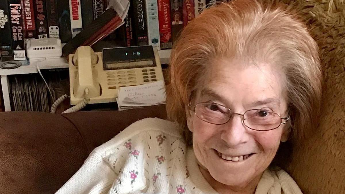 A elderly woman smiling at the camera. She has thin black frame glasses and short blonde/light brown hair. She is wearing a white top with small flowers on them. There is a landline telephone behind her and she is sitting on the sofa