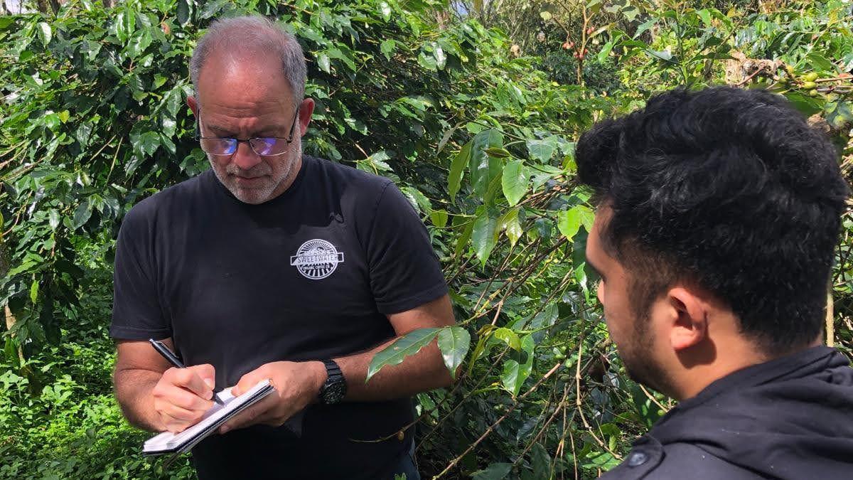 Pomeroy is wearing a black t-shirt and writing in a notebook with a black pen among green foliage, with the back of the head of a farmer in the foreground