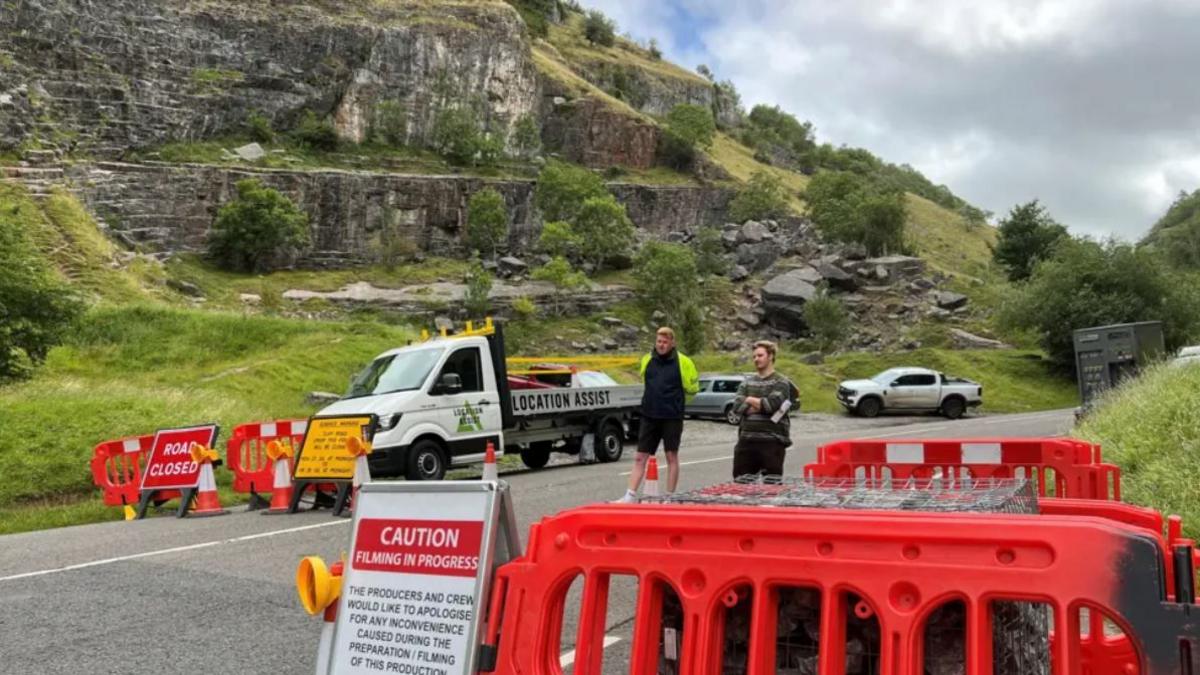 A winding road in Cheddar Gorge which has been closed with plastic fencing and orange cones. There is an A-board sign on the side of the road which says Caution, filming in progress. There are two men stood in the middle of the road, one with his arms crossed and the other wearing a hi-vis vest.