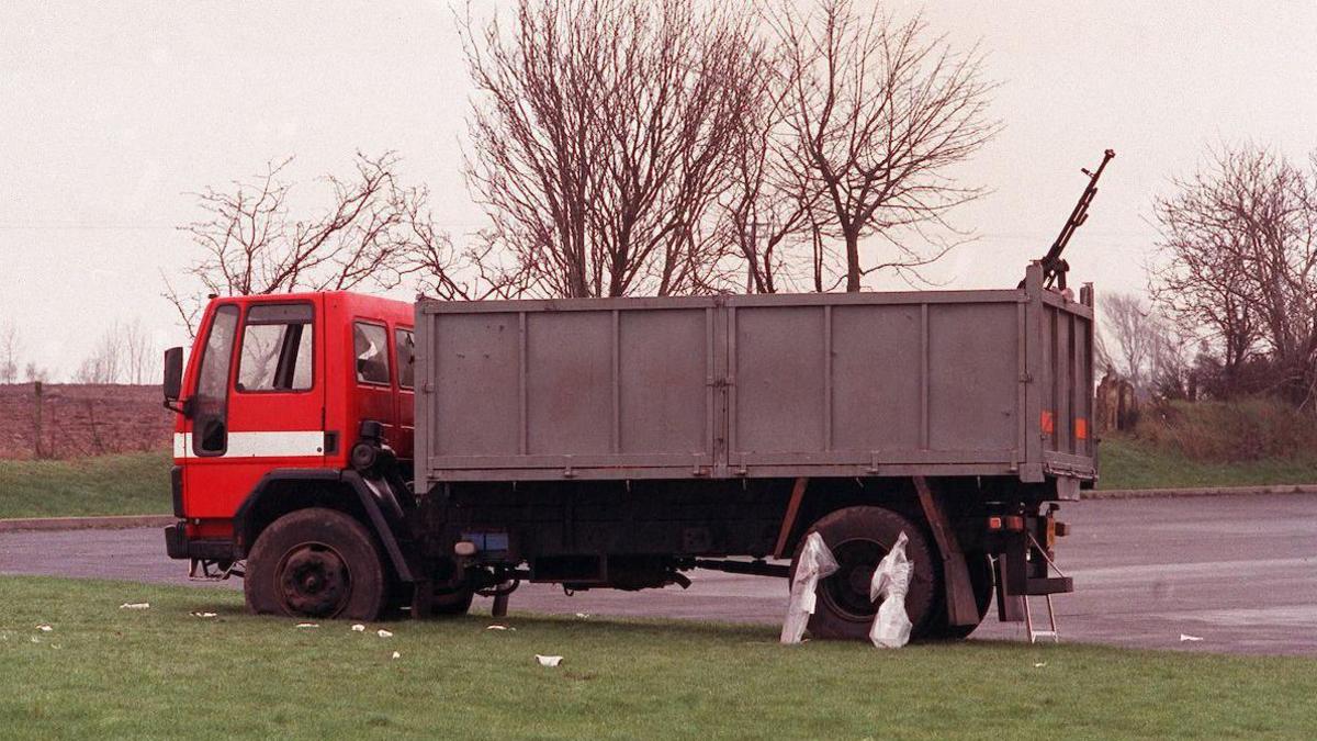 A red lorry with a grey carriage. Attached to the tailgate of the lorry is a heavy black machine gun, pointed to the sky. The lorry is sitting on a patch of grass beside a carpark.