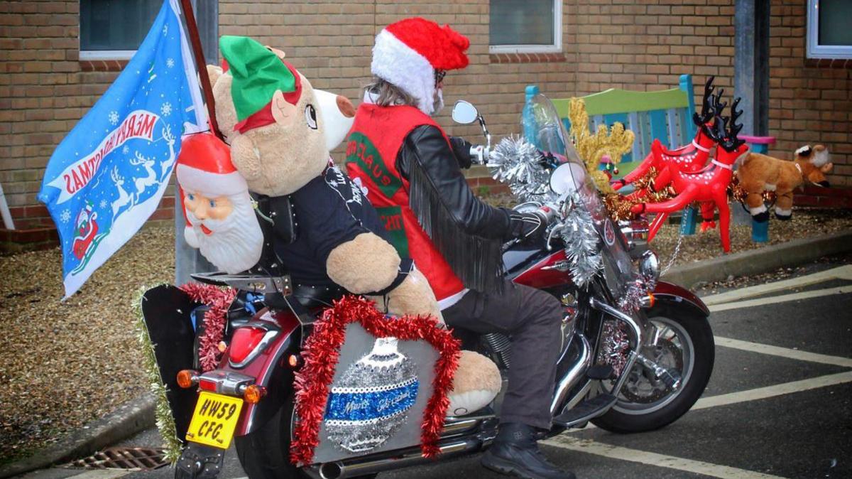 Buster Spicer on his bike in front of the hospital. He's facing away from the camera. He has a big plush bear on the bike behind him. There is also a blue flag with Santa's sleigh and the inscription "Merry Christmas".