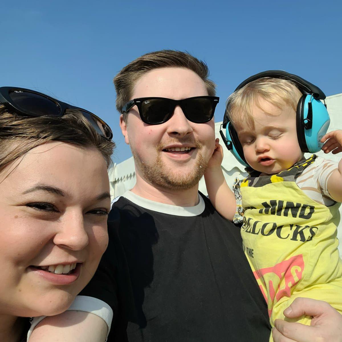 A man in a black t-shirt takes a selfie whilst holding a child wearing blue ear defenders, to the other side is a woman whose face is just above his arm