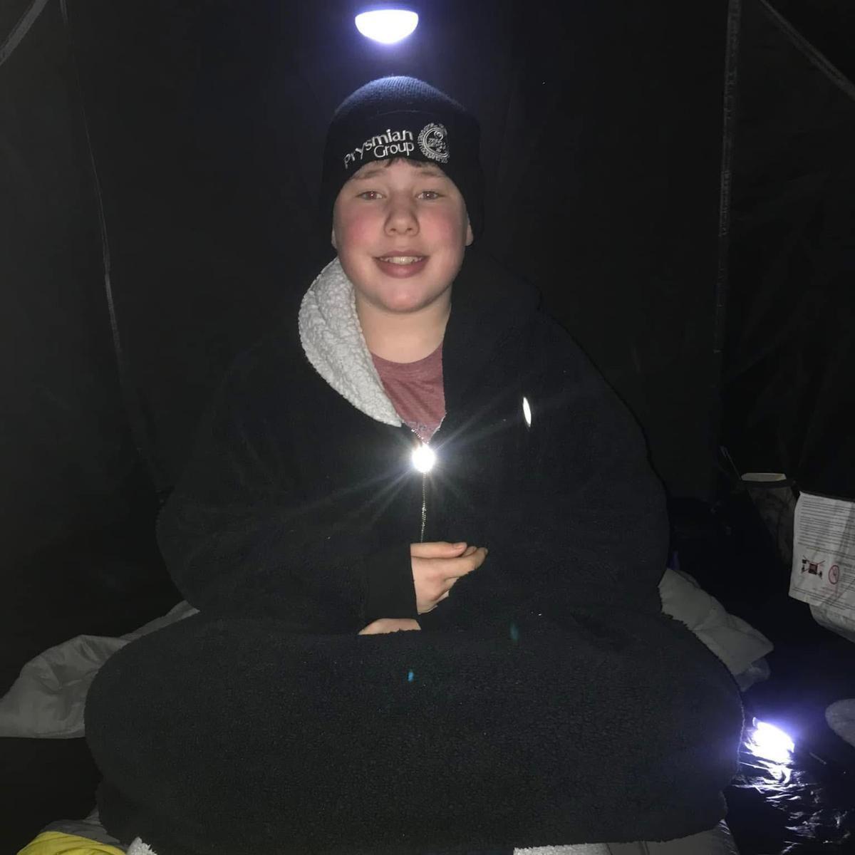 A boy in black clothes and a black hat, sits cross-legged in a tent while the light from a torch can be seen on his chest.