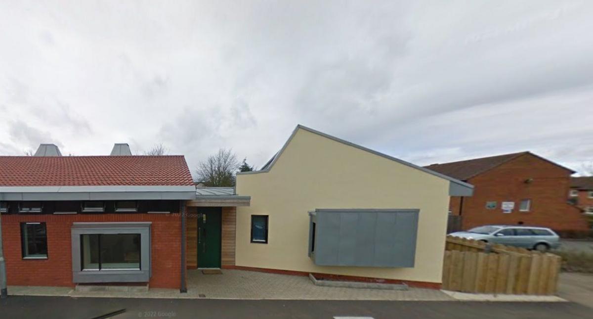 A single-story brick-built house, with a brick coloured section on the left and painted cream-coloured section on the right. There are two large picture windows - one of which has a metal shield over it.