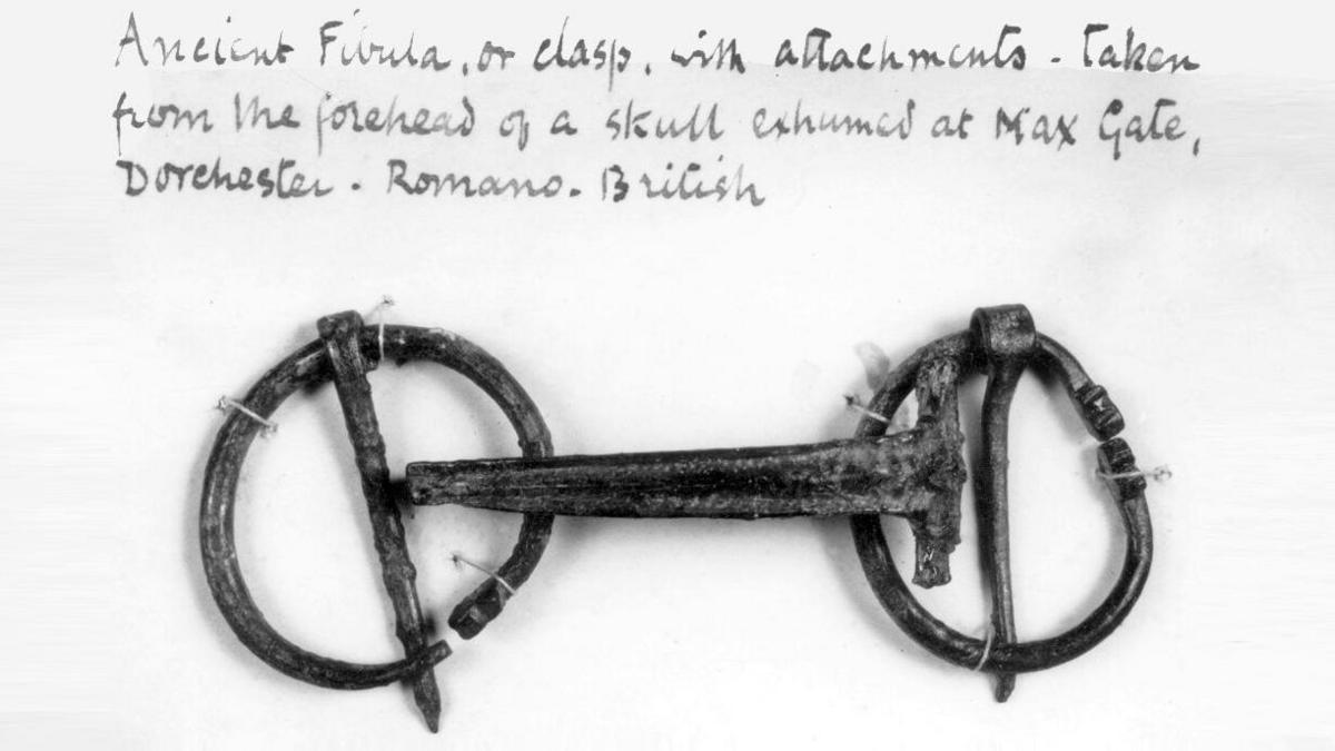 A black and white photo of a bent clasp with two circular ends arranged below a handwritten note from Thomas Hardy which reads: Ancient fibula, or clasp, with attachments - taken from the forehead of a skull exhumed at Max Gate, Dorchester - Romano-British