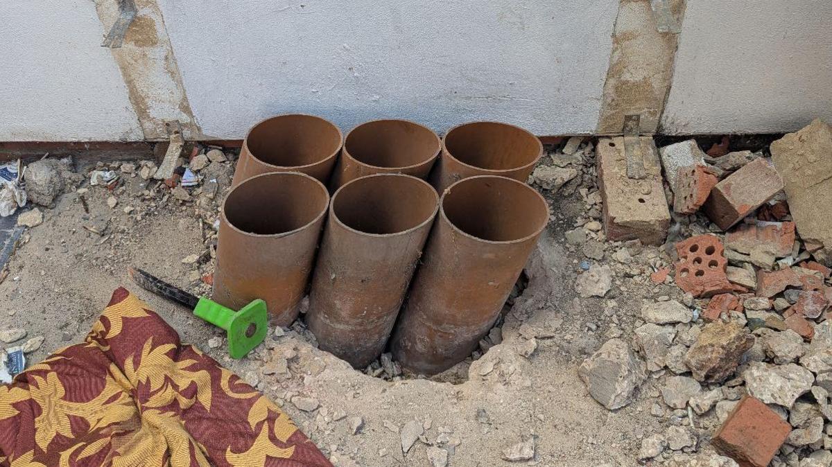Six brown pipes can be seen sticking out of some concrete. There is rubble on the ground nearby and a tool which was used to chisel away at the concrete