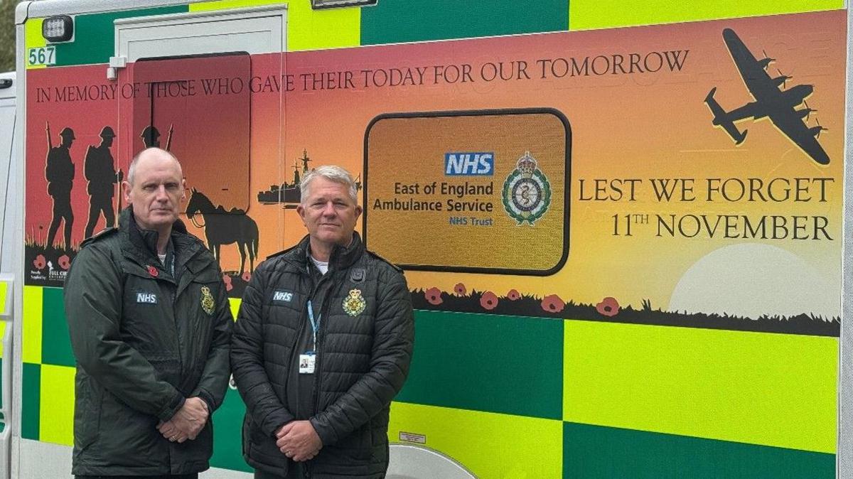 Two men are stood on the left of the image looking towards the camera. Behind them is an ambulance which has a design on it for Remembrance Day, including a plane, red poppies and soldiers.