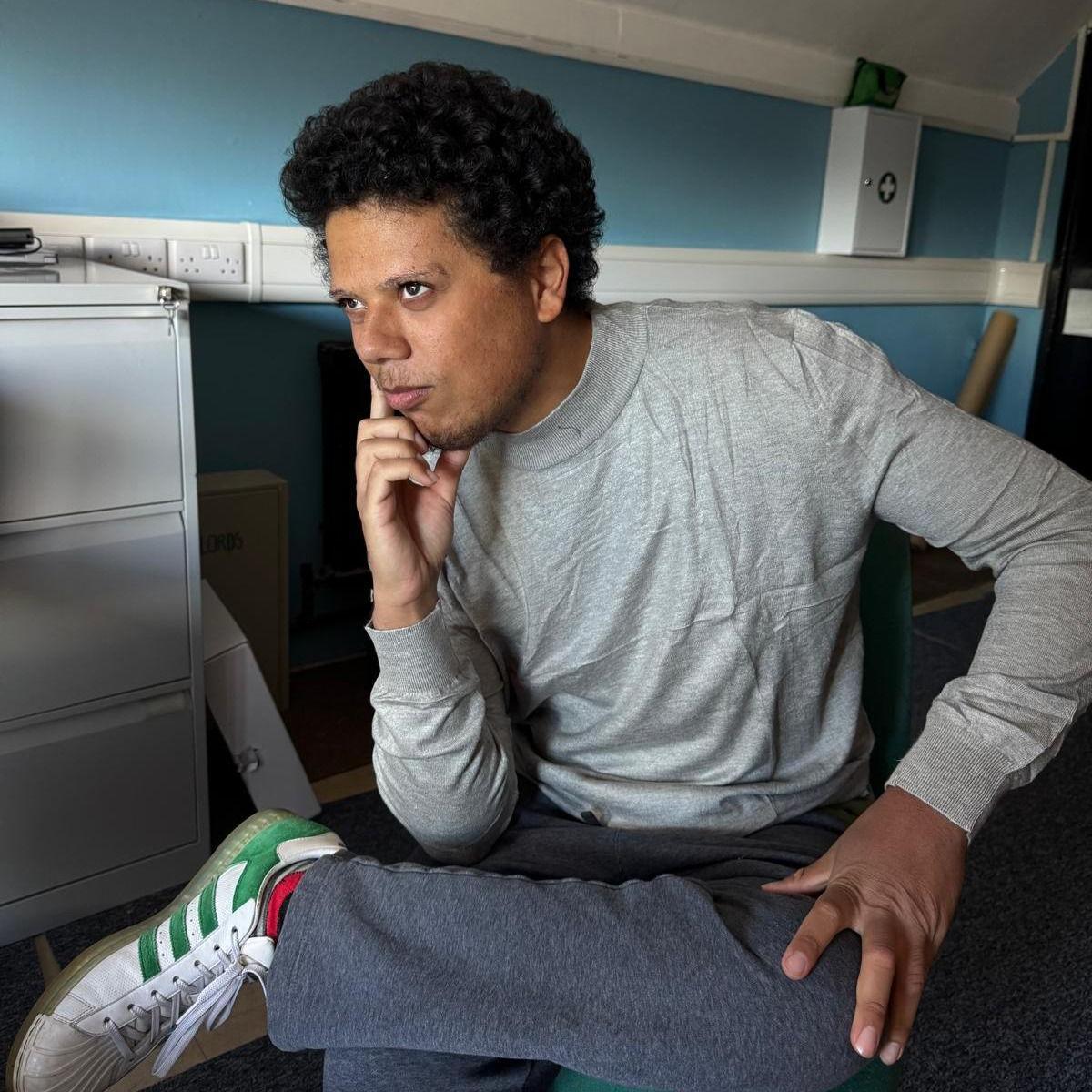 A man with dark, curly hair and a short beard is wearing a grey sweater with blue trousers and white trainers as he sits on a chair with his left leg propped on his right knee, leaning his head on his right hand. He is sat in an office with blue walls and a white filing cabinet seen behind him.