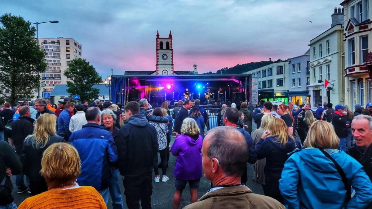 People in colourful raincoats and jumpers standing in front of a brightly lit stage in a built up area. There is a pink hue in part of the cloudy sky as the sun sets in the background.