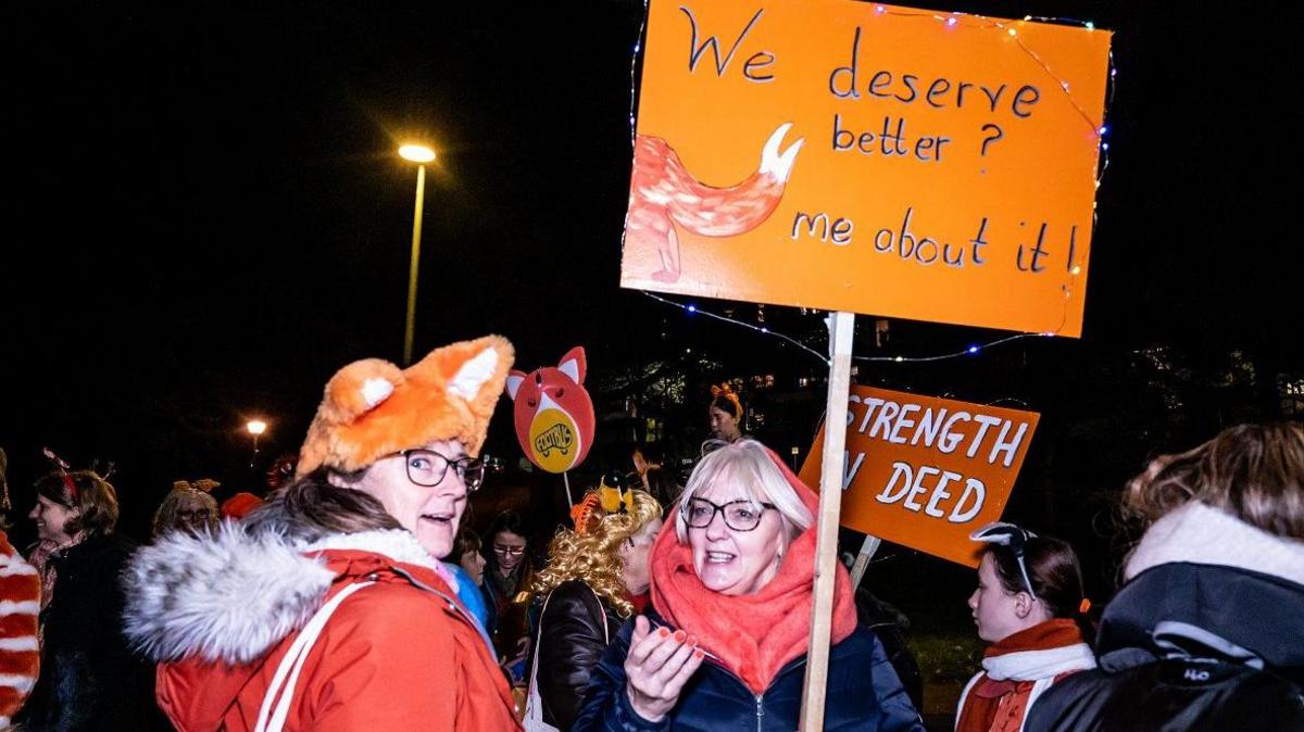 Two women who appear to be mid-conversation with one another, wearing orange. One of them is wearing a fox hat and the other is holding up a sign saying "We deserve better? Tell me about it". It's night time and there are other people behind them.