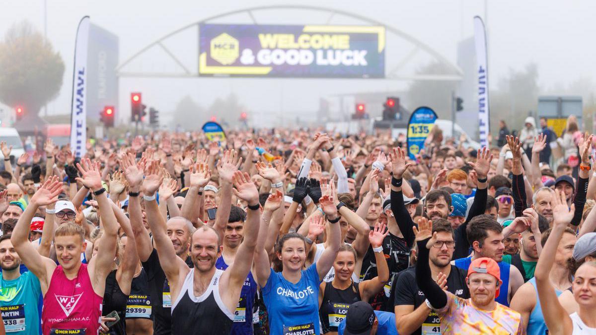 Thousands of runners mass at the start line raising their arms and smiling with an arch above them int he background that has a sign saying "Welcome and good luck". There is fog above.