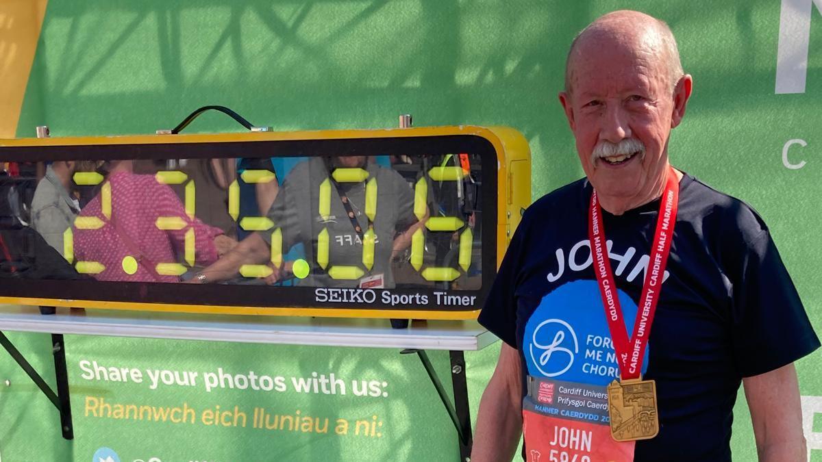 John ar ddiwedd hanner marathon o flaen cloc gyda'i amser