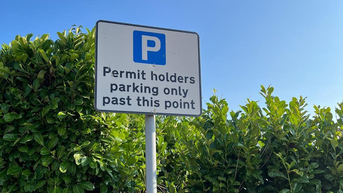 A sign has a blue 'P' parking sign onit and reads 'Permit holders parking only past this point'. It is standing on a pole in front of a green hedge and blue sky.