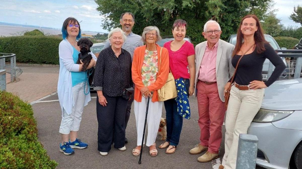 Seven people standing together in a car park. The sea is visible in the background.