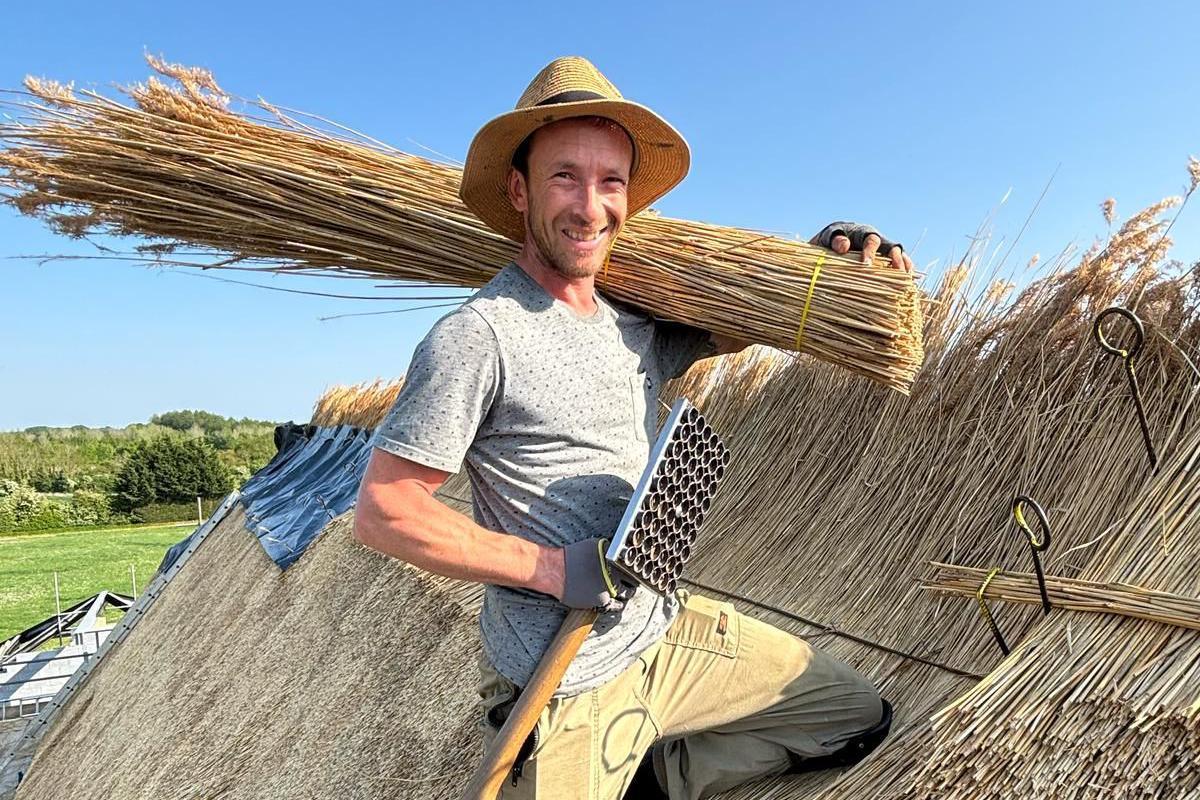 Luke is kneeling on a thatched roof, with long straw thatch tied together placed over his left shoulder. He is wearing a sun hat, grey t-shirt and tan-coloured combat trousers. There are blue skies above.