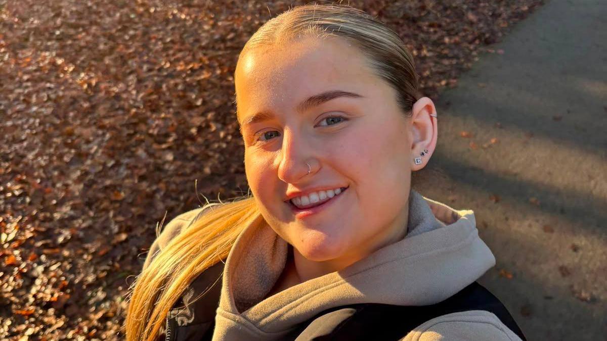 Picture of a young woman smiling for a selfie in the woods. She has blonde hair slicked back in a ponytail and is wearing a stone coloured hoodie with a black body warmer.