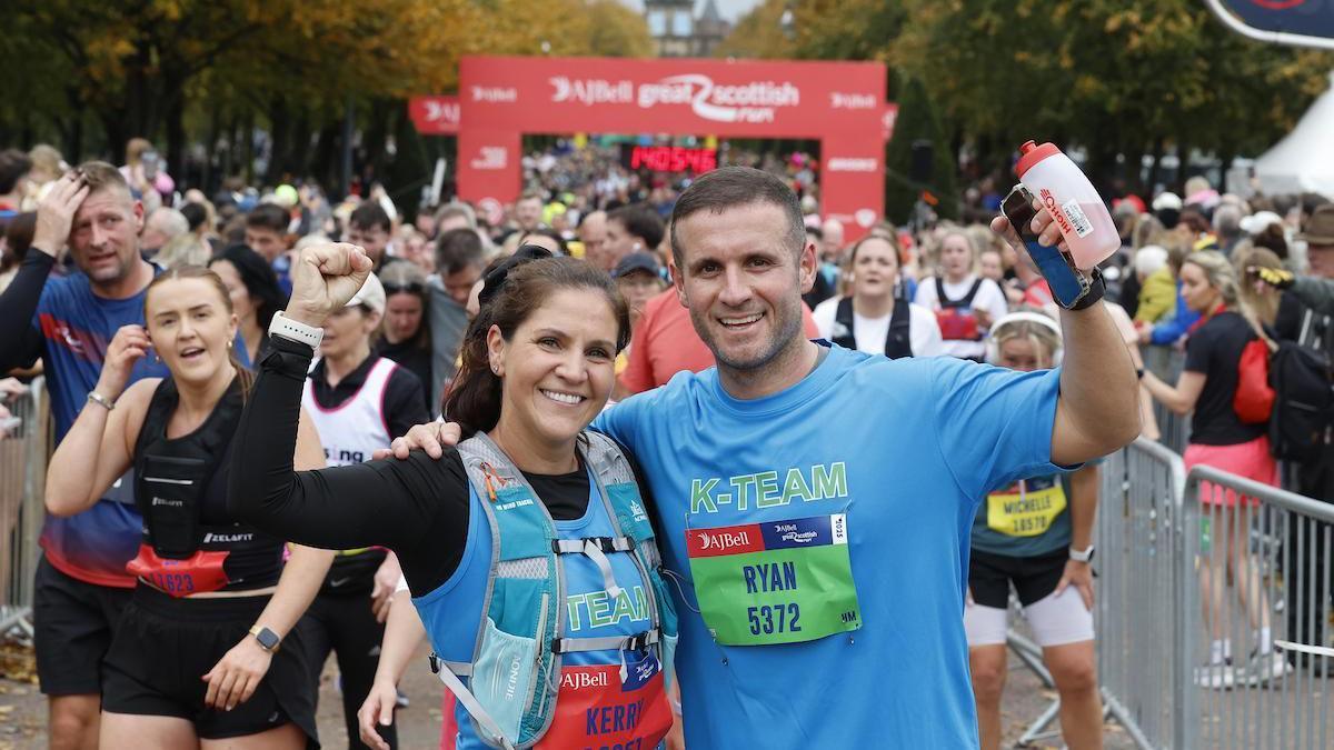 A man and woman both wearing blue tops, with one arm round each other and the other up in the arm celebrating as they cross the finish line at the AJ Bell Great Scottish Run.