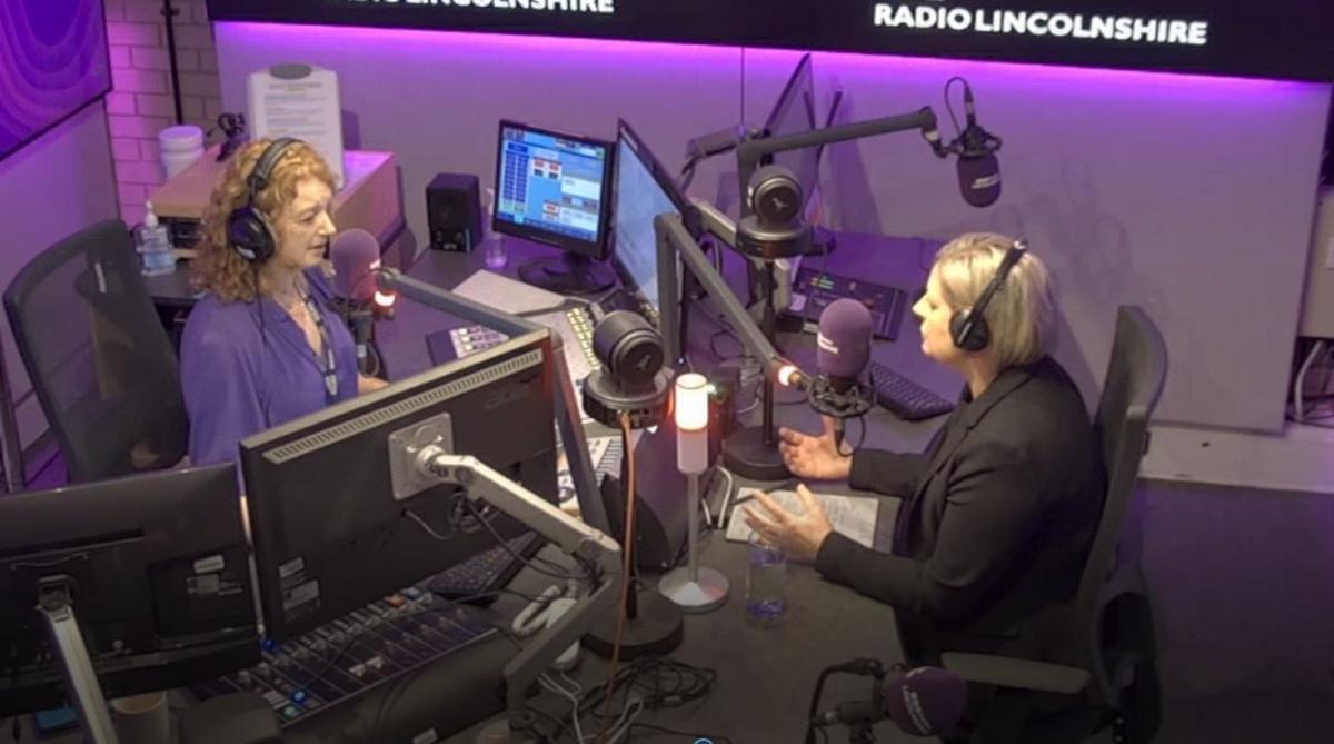 Professor Karen Dunderdale and BBC presenter Frances Finn sitting in a radio studio. They are surrounded by computer screens and microphones with a mixing desk in front of them. The walls of the studio are bathed in purple light with a black Radio Lincolnshire logo on them. Frances has ginger curly hair and is wearing a purple shirt