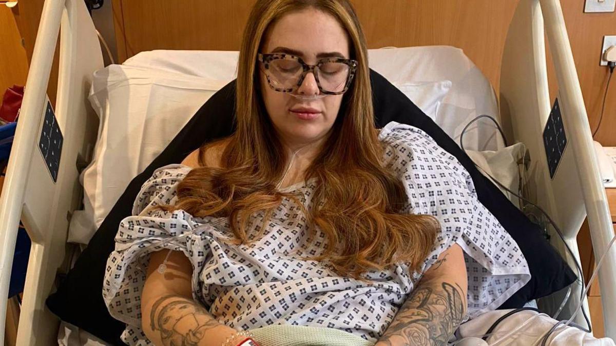 A woman in a hospital gown, Gabriella Pearson, looks down with her eyes closed as she lies on a hospital bed wearing a hospital gown.