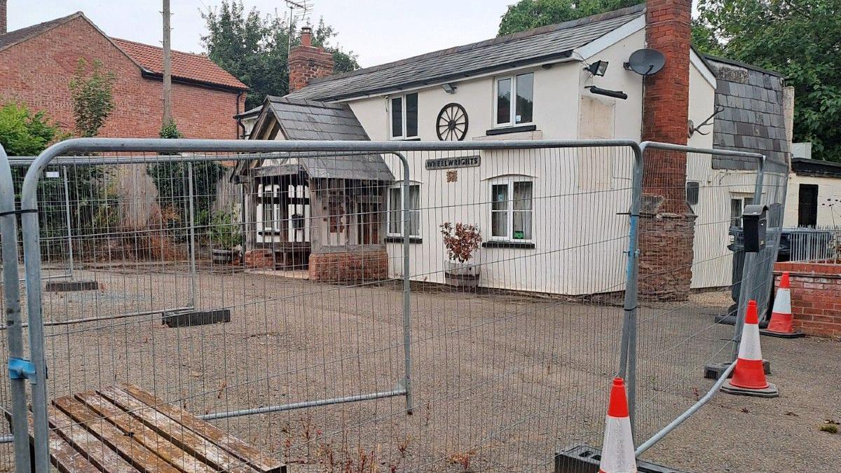 A white-coloured and vacant village pub behind a steel fence which has several cones next to it. Another house can be seen on the other side, with trees behind the buildings.
