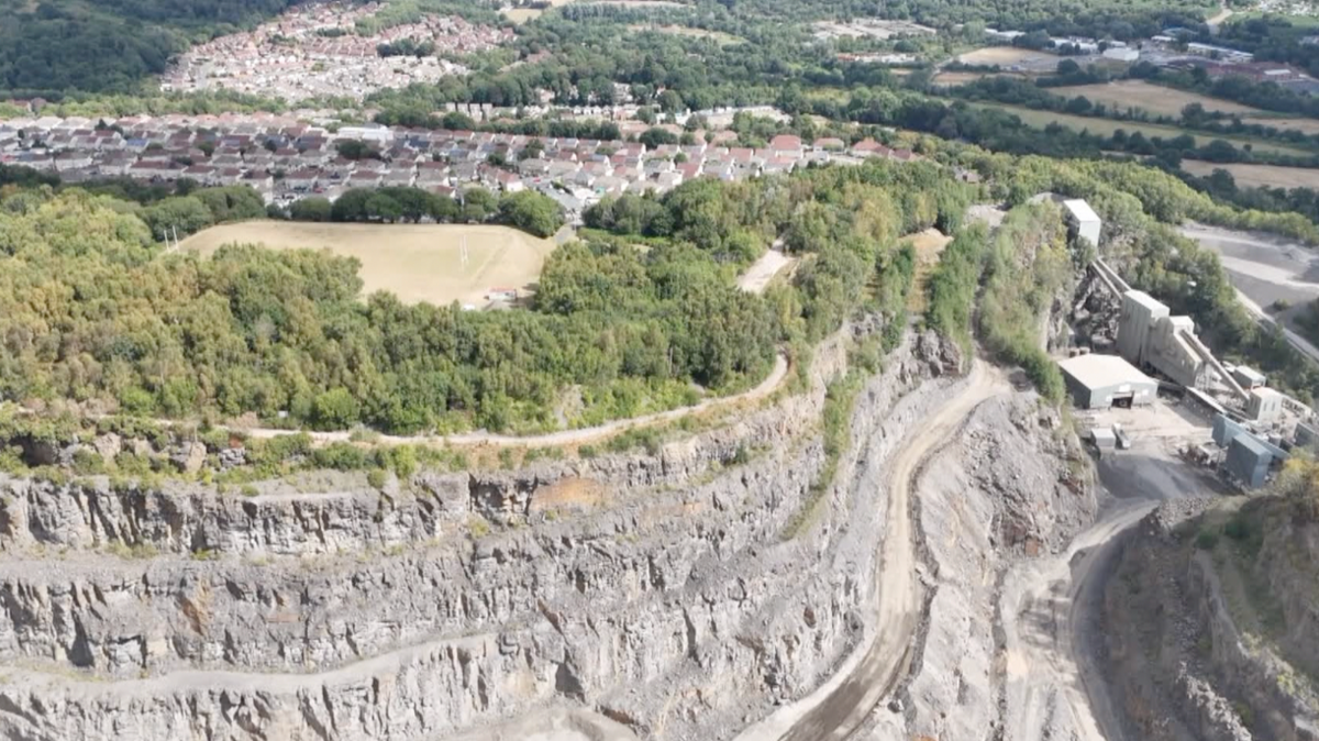 The rock face of the quarry in the foreground. In the backdrop there are green trees, arugby pitch and then rooftops of a nearby village alongside other yellow and green fields