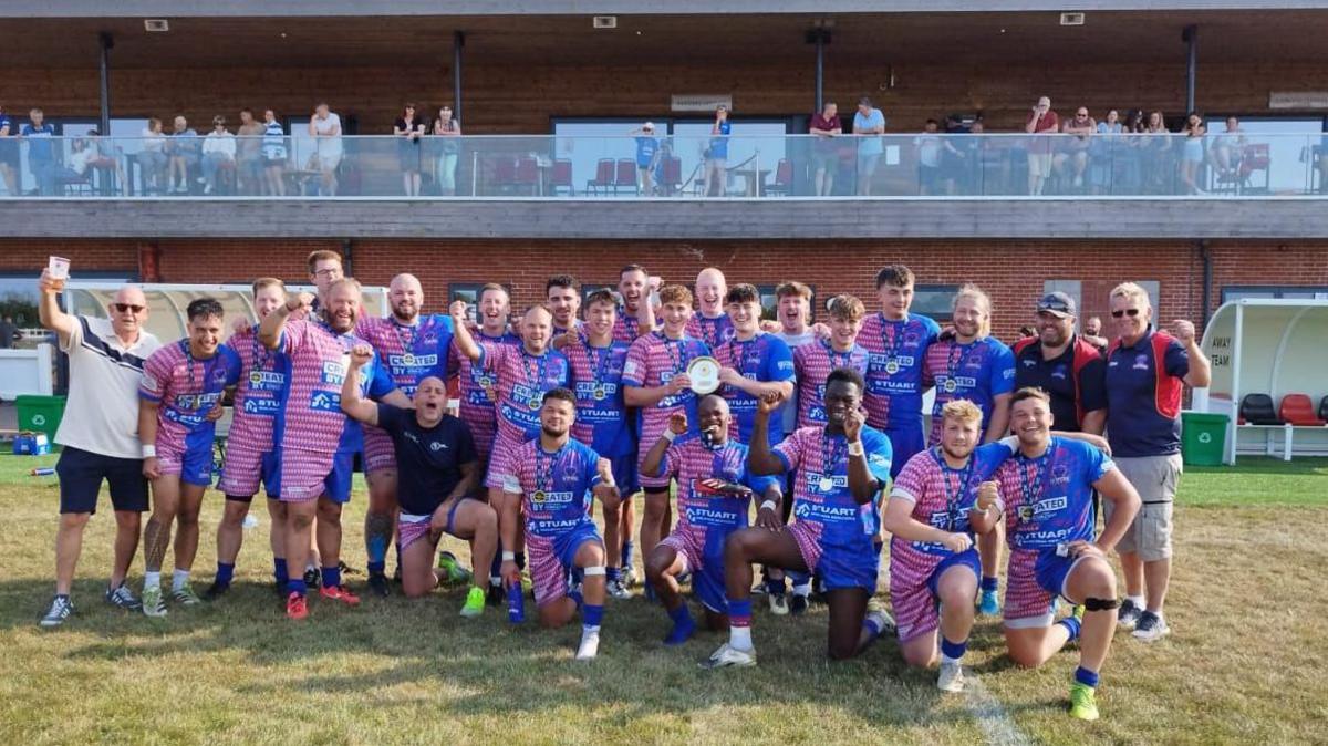 The Anglian Vipers squad pose for a team photo pitchside with the East Rugby League trophy as fans, friends and family look on from a balcony behind them