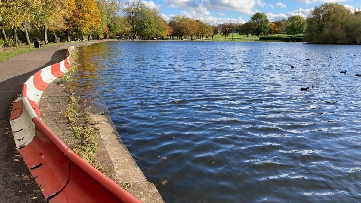 Pontefract Park lake showing the section which needs repair, cordoned off with a red and white metal barrier