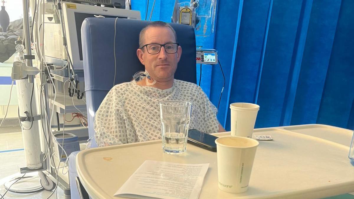 Jonathan in a hospital chair surrounded by wires and machines. There is a table in front of him with cups of coffee and a glass of water. The man has very short hair and is wearing black frame glasses. He is in a hospital gown and has a cannula in his neck.