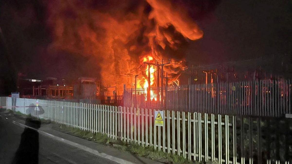 A general view of an electrical substation fire in Hayes, West London. The fire is behind a metal fence. There is another smaller fence in front. Both are grey. The fire is orange and is around electrical pylons.