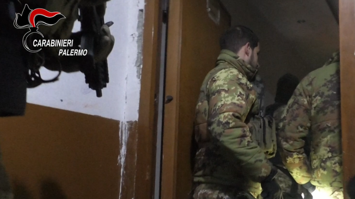 Two uniformed officers of the Carabinieri wearing jackets with a camouflage print stand in a doorway while a colleague behind them holds a gun