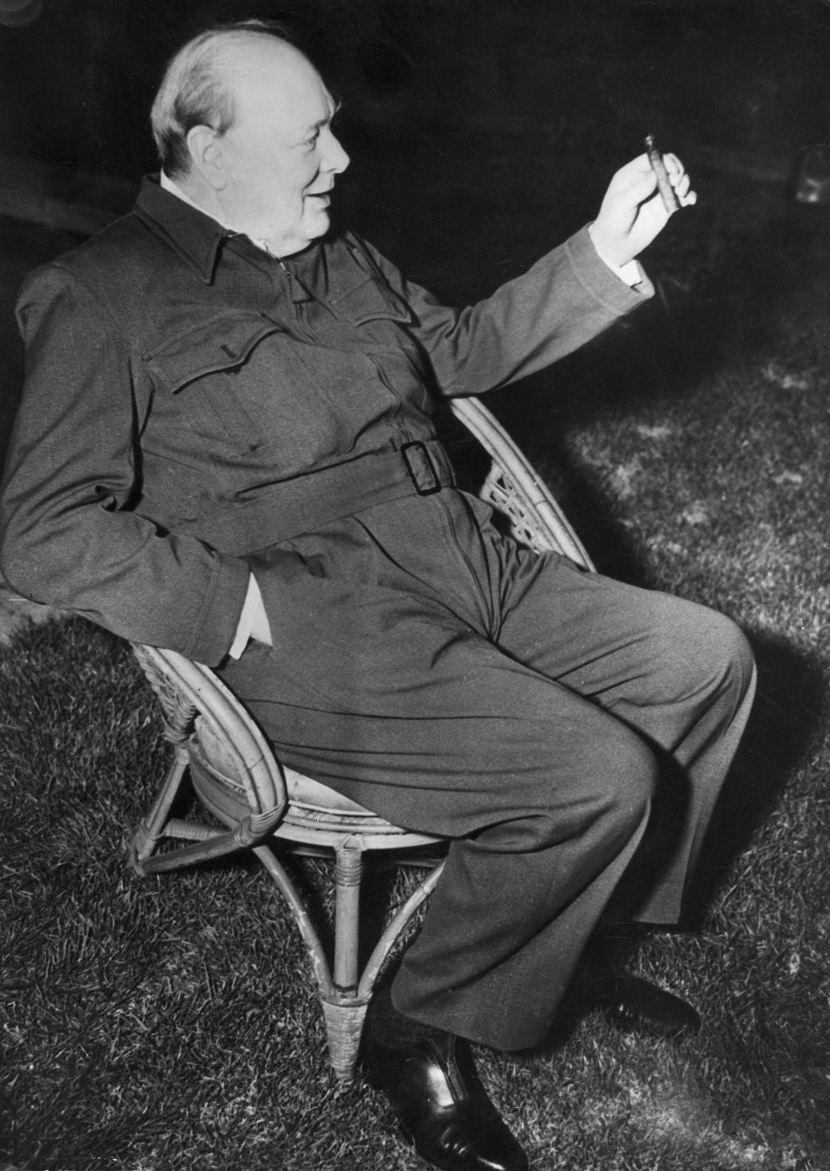 A black and white photo of a seated Winston Churchill clutching a cigar and wearing a suit that resembles a jump suit or modern-day onesie with vest pockets and a belt