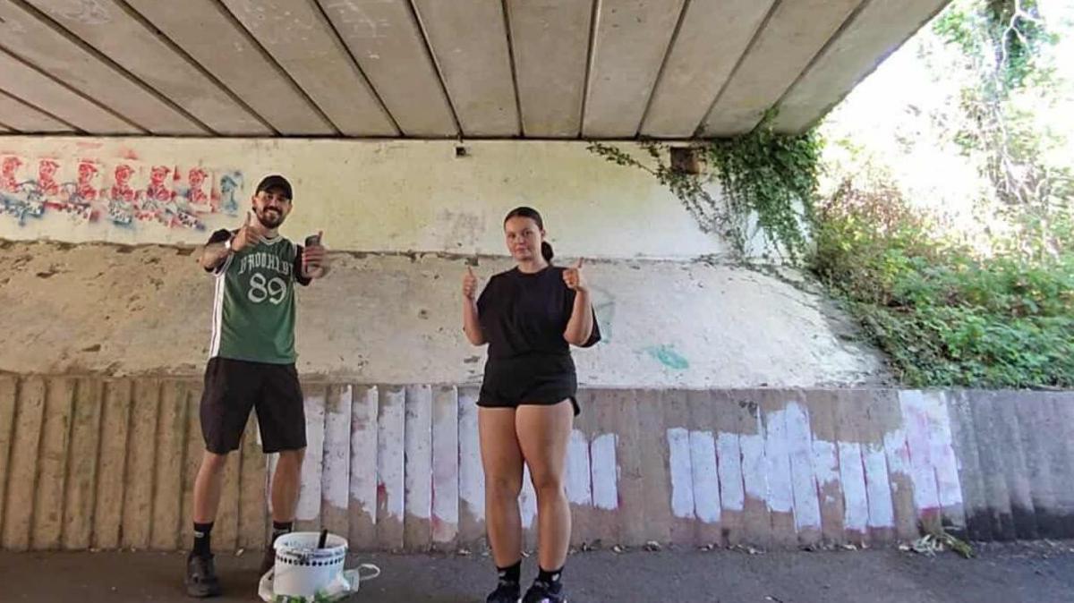 Photograph of a young couple stood inside a subway walk way. A girl (right) wears a black t-shirt and black shorts and holds both her thumbs up. While the young man (left) wears black shorts, black shoes and a green baseball vest and holds his thumbs up to the camera. A bucket of white paint can be seen infront of them, and patches of white paint can be seen on the wall behind them.