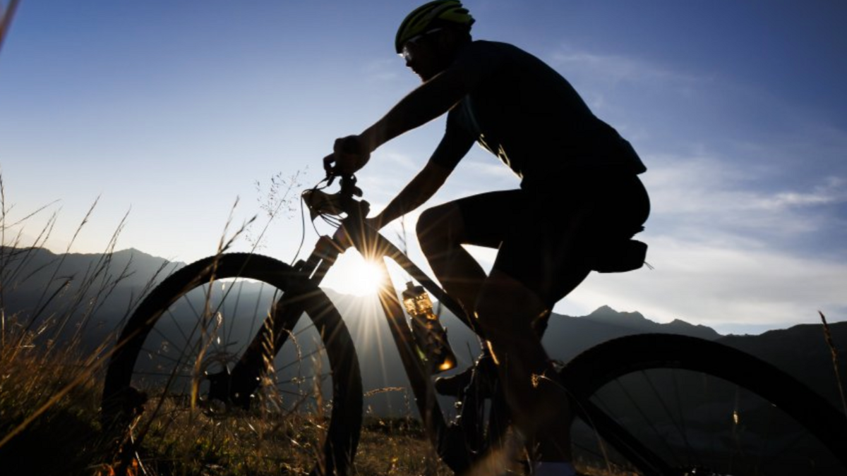 Mountain bike cyclist on off-road track with hills or mountains silhouetted in the background with the sun shining over the top of the hill. The cyclist is also a silhouette.