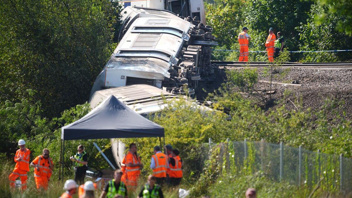 Several derailed train carriages lie on their sides down a railway embankment near Lewes in East Sussex. Rail workers and investigators in orange high-visibility clothing stand beside the track and in a field below, where a temporary canopy has been set up as part of the investigation.