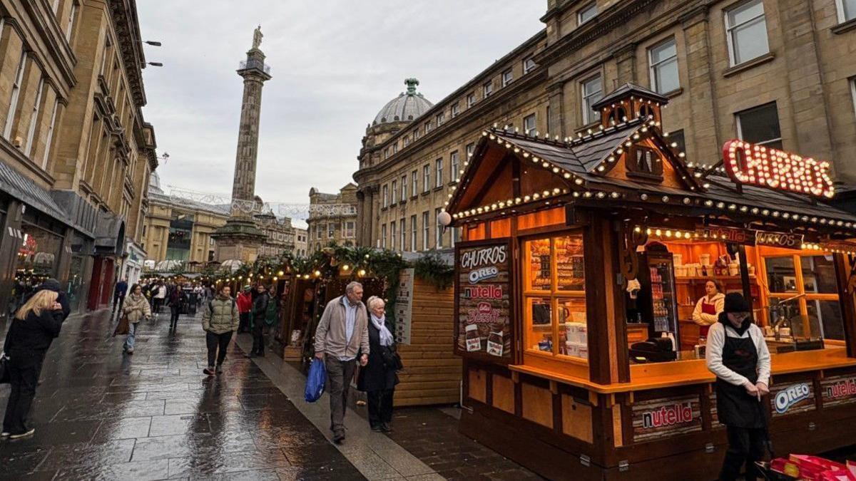 A number of wooden stalls in this year's Christmas Market, including a churros stall in the foreground on the right-hand side. Several people are walking along the street. The pavement is wet as it is raining. A number of ornate stone buildings can be seen on either side with Grey's Monument - a stone column - rising in the background.