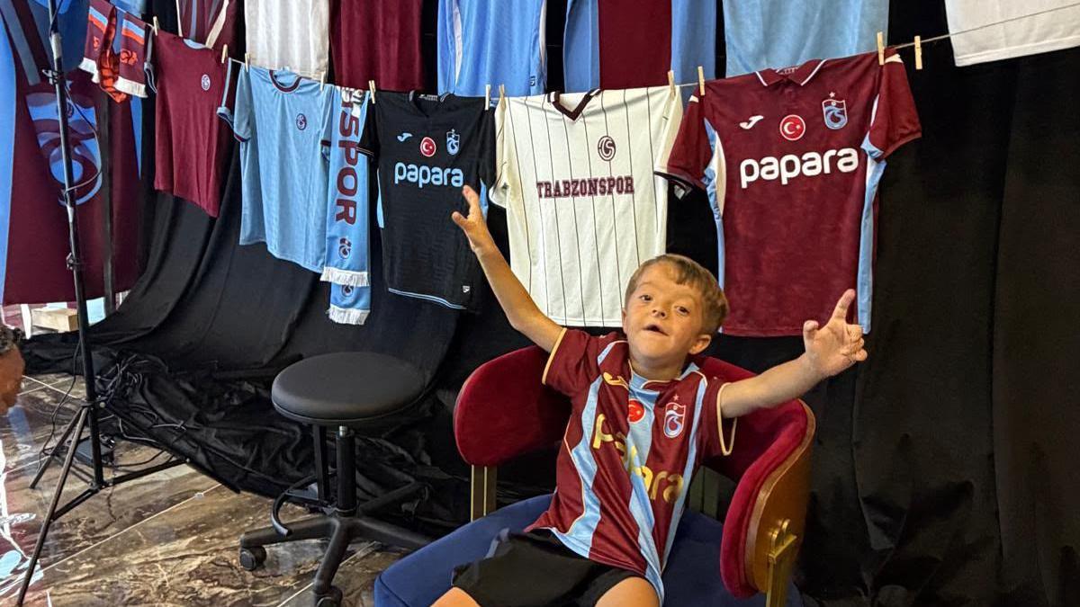 Isaac raises his arms while sitting in front of a display of Trabzonspor shirts and scarves.