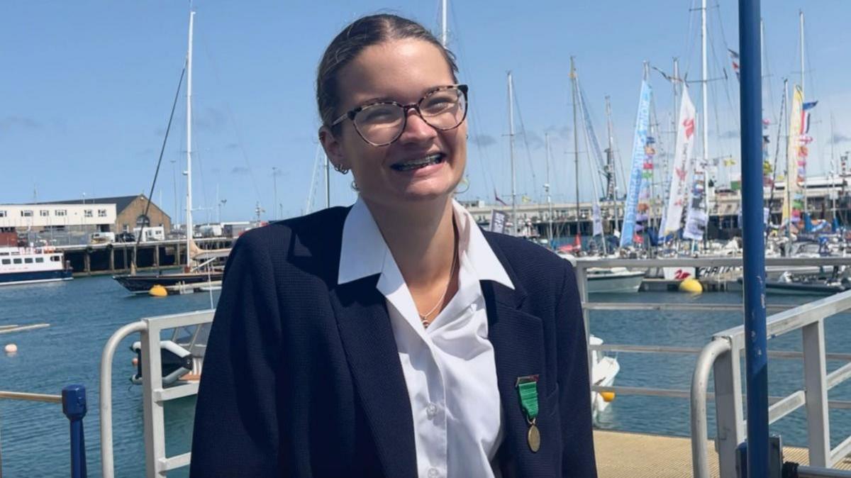 Neave Chatting-Tonks - girl with brown hair in a ponytail, tortoise shell glasses brown and black, big smile with braces, wearing a white shirt under a navy blazer with a medal on the left lapel, background of the sea and a row of different boats on a clear sky day