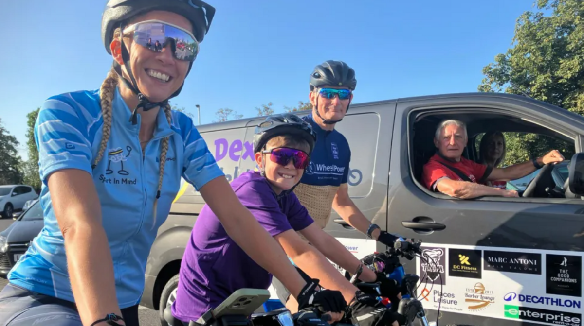 From left to right - Dexter's mother Sam, Dexter and coach Dave Covey as smiling as they are prepared to set off. All three are on their bikes. Sam wears a blue top for Sport in Mind, Dexter wears a purple T-shirt for Falconi Hope, and Mr Covey wears a dark blue and yellow top for WheelPower. A driver in a van with Dexter's name on it is next to them. A woman can be seen in the passenger seat. It's a sunny day.