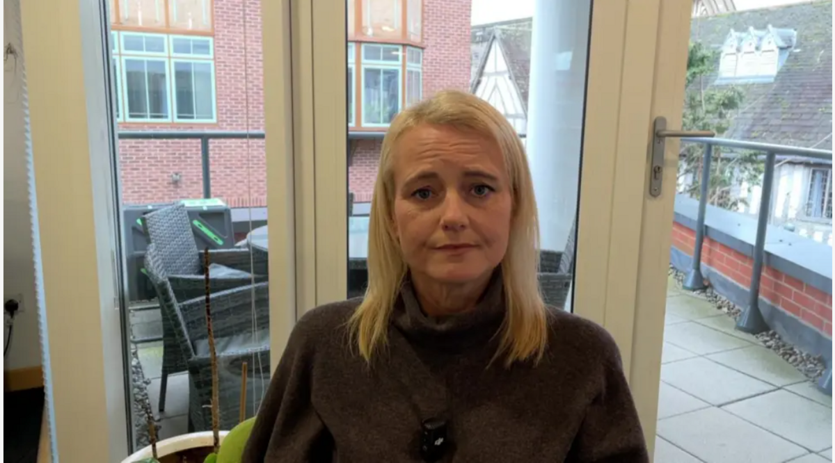 Justine Carter sitting in her office with a window and patio area behind. She has shoulder length blonde hair and is wearing a brown jumper