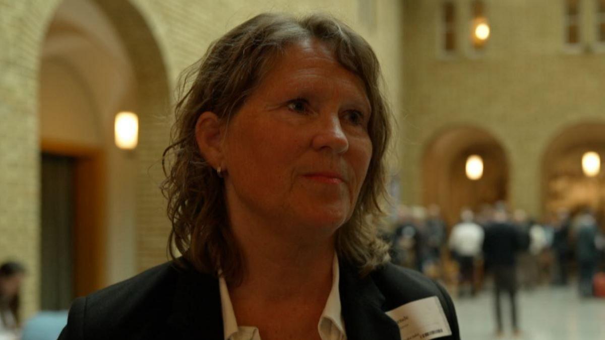 Merete in a large hallway. She has shoulder length wavy brown hair and is wearing a white shite and dark suit jacket