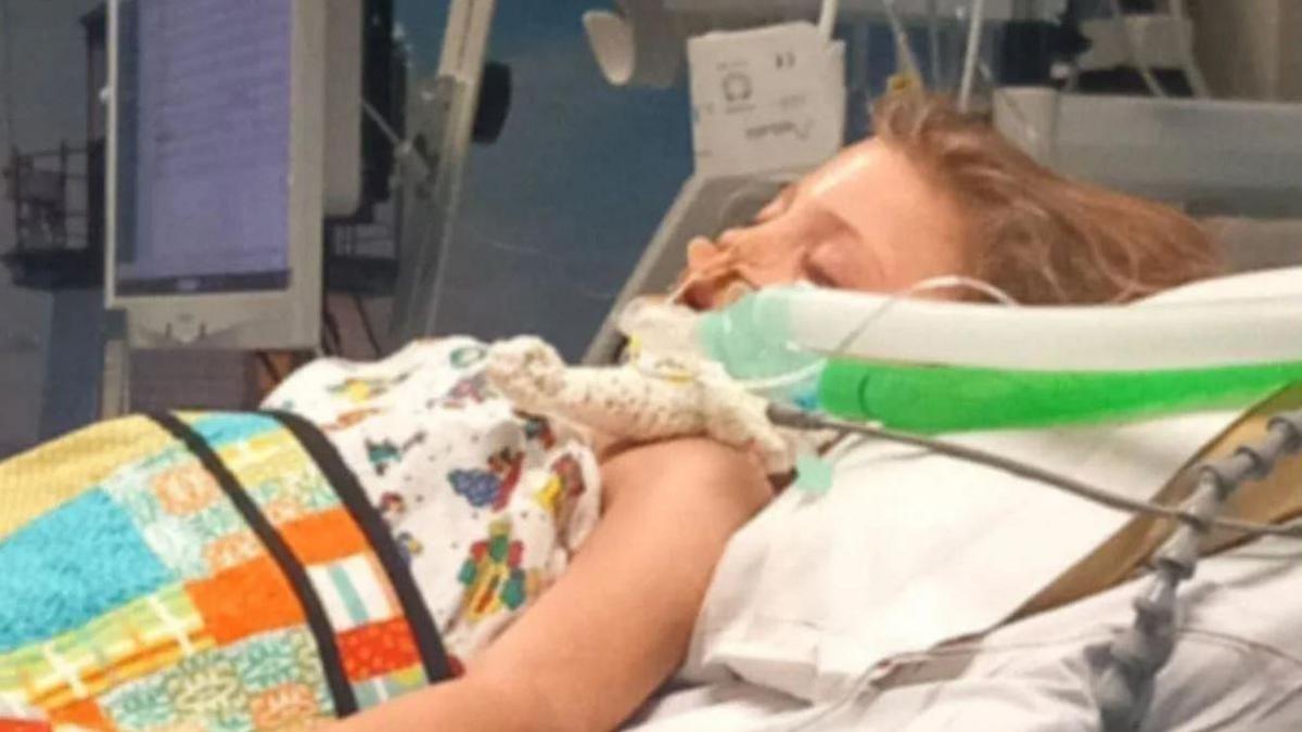 A little girl with blond hair, lying in a hospital bed underneath patterned blankets. There are tubes going up her nose and down her throat, and she has her eyes closed. 