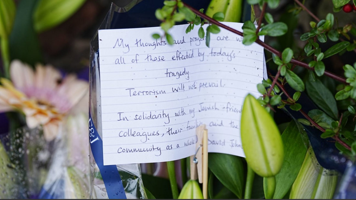 A handwritten note among flowers on lined card. Not all words are visible but it starts: "My thoughts and prayers are with all of those affected by today's tragedy. Terrorism will not prevail."