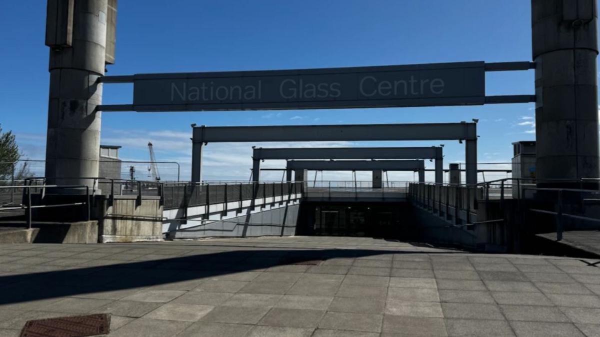 The entrance to the National Glass Centre in Sunderland with two pillars on either side of a paved path leading down underground. A metal banned over the path reads National Glass Centre.