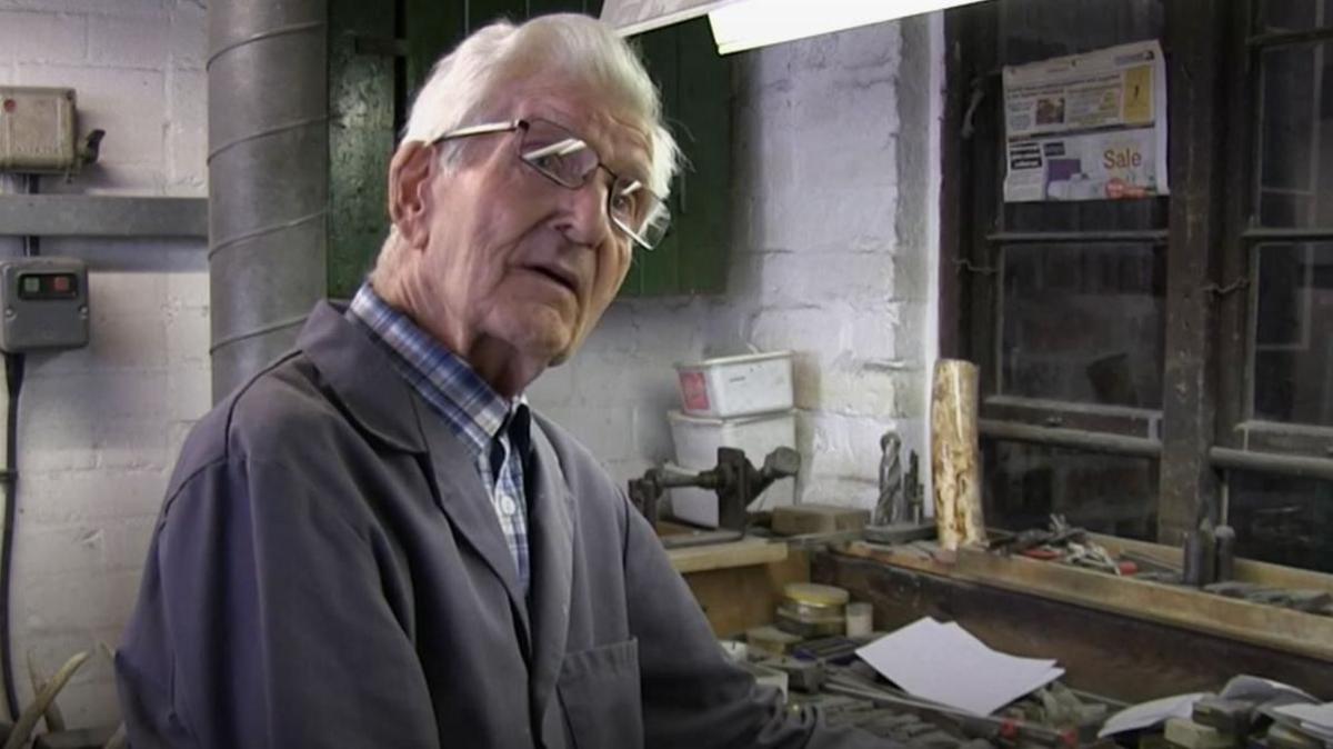 A man, Stan Shaw, with grey hair, wearing glasses and blue overalls in a workshop
