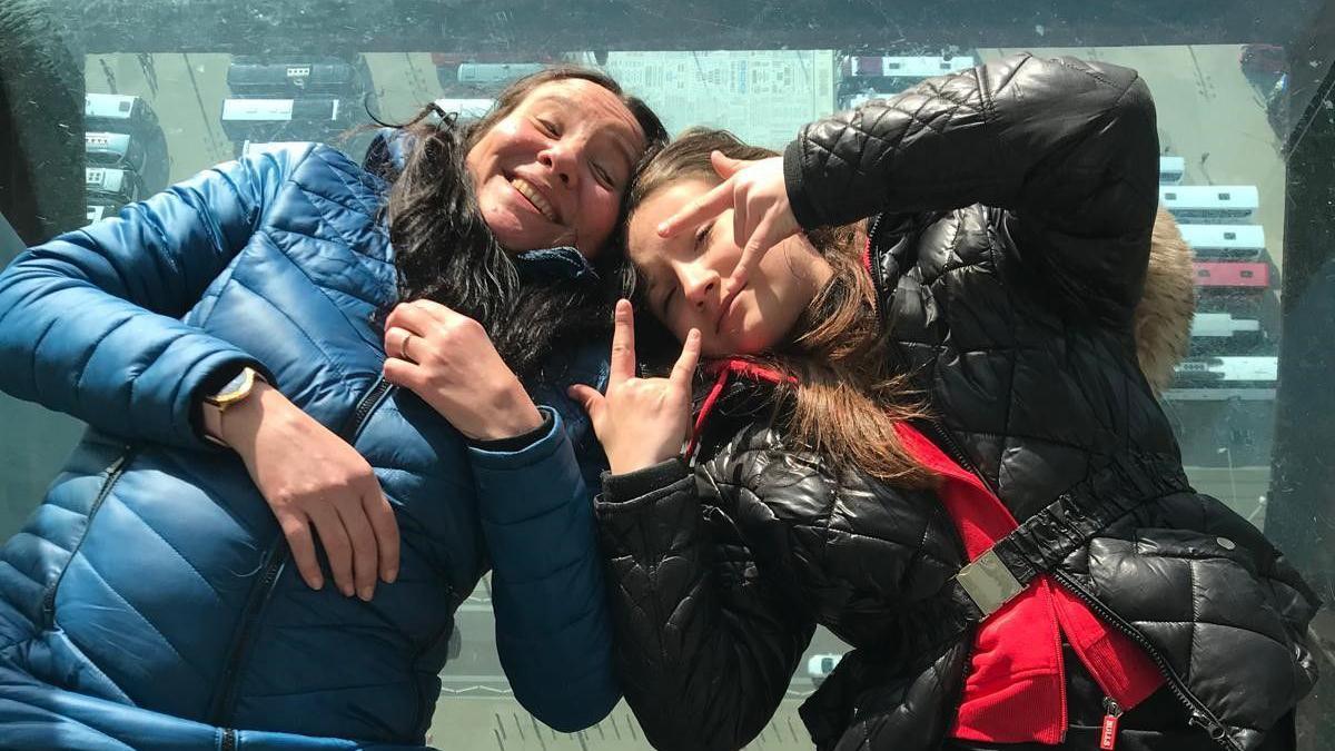 A mother poses with her teenage daughter. They are leaning in towards each other, with heads touching and facing the camera. The mother is smiling and wearing a blue coat while the daughter makes hand signals at the camera in her black coat. 