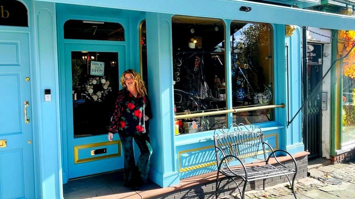 Jane McNally is wearing a black, red and green floral shirt and green trousers. She is standing in the doorway of her shop which is painted light blue with a gold trim.