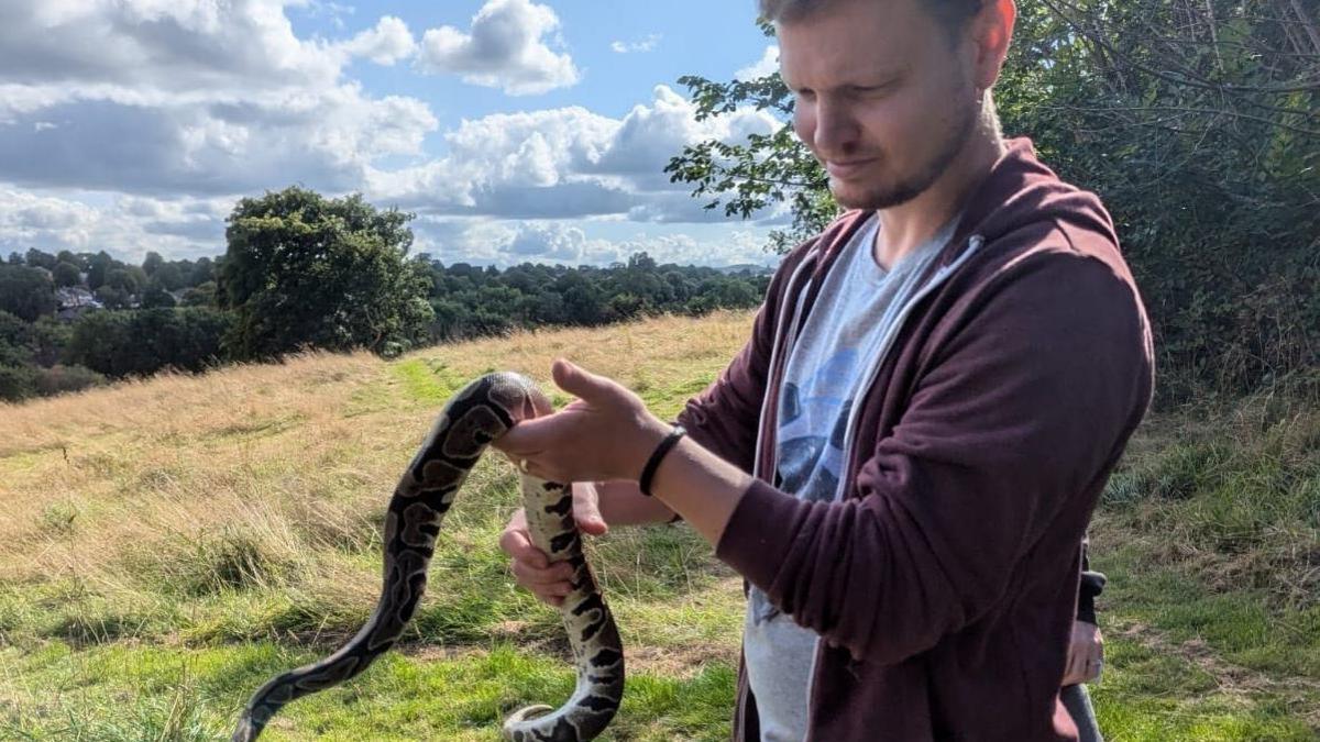 Tom Gallant, whose family found the python, stands in the park with the snake in two hands - he has a beard and is wearing a burgundy zipped top and blue T-shirt