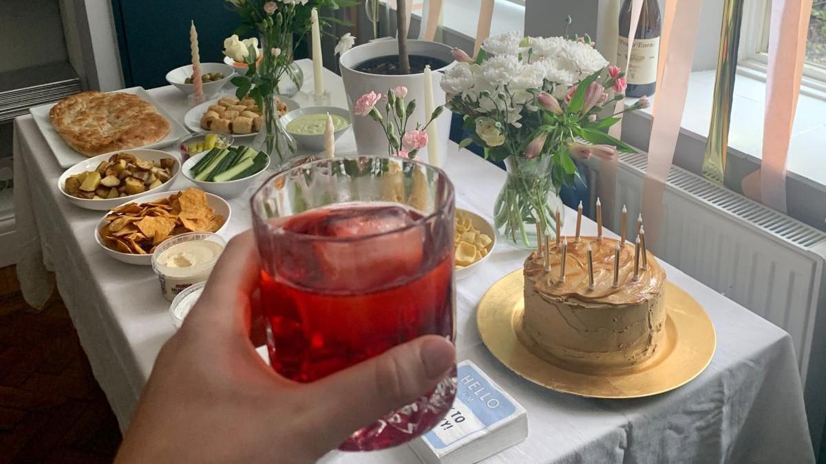 Jess Warren holds a glass of red liquid with ice, in front of a table. On the table is a birthday cake, crisps, bread and vegetables. There are four vases of flowers.