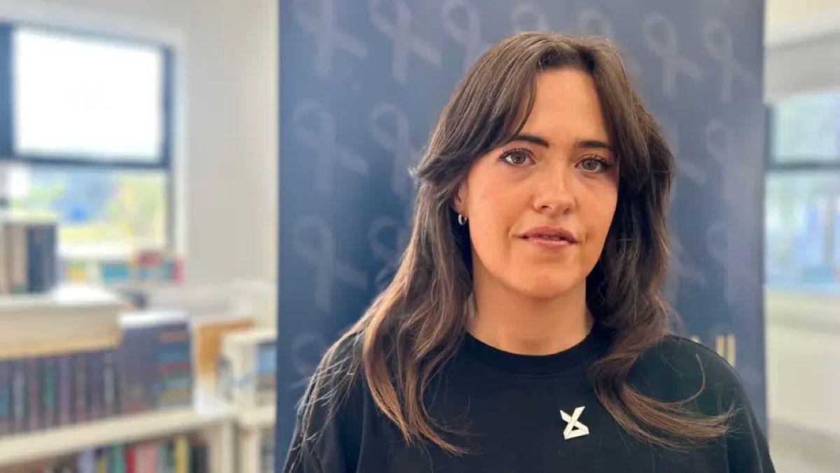 Tahnee McCorry has long brown hair, wearing a black top. She is looking at the camera. There is a dark blue poster behind her with light blue ribbons on it. There is a bookcase behind the poster.