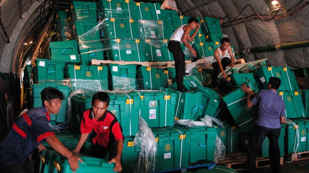 A group of men help to load green boxes out of the hold of an aircraft. The boxes are stacked up to six high and are wrapped in cling film and cable ties.